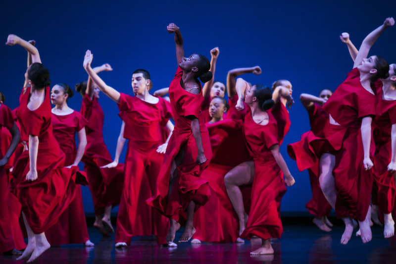 A group of young dancers in bright red shirts and long skirts lift their fists in the air. 